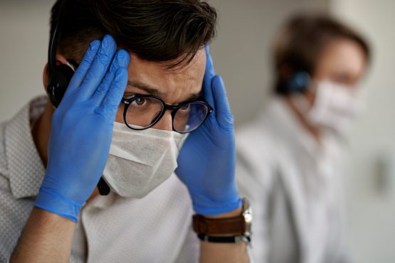 Customer service representative feeling tired and holding his head in pain while working at call center during coronavirus epidemic.