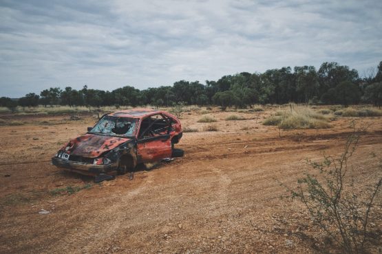 An old broken up car in a dry grass field with trees in the background